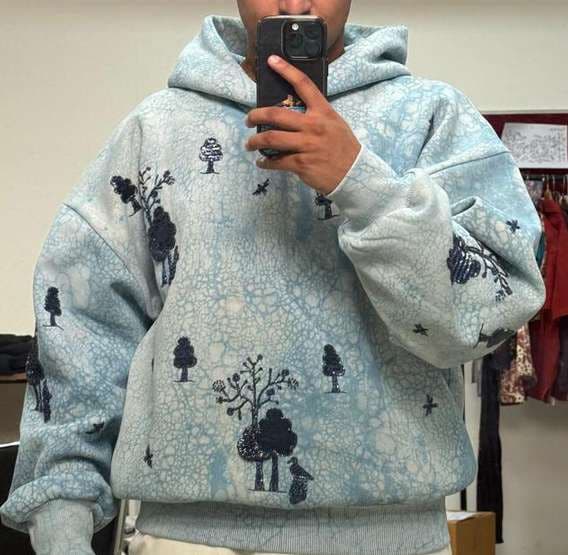 A mirror photo of a pale blue hoodie with a mottled, crackled surface and printed tree motifs.