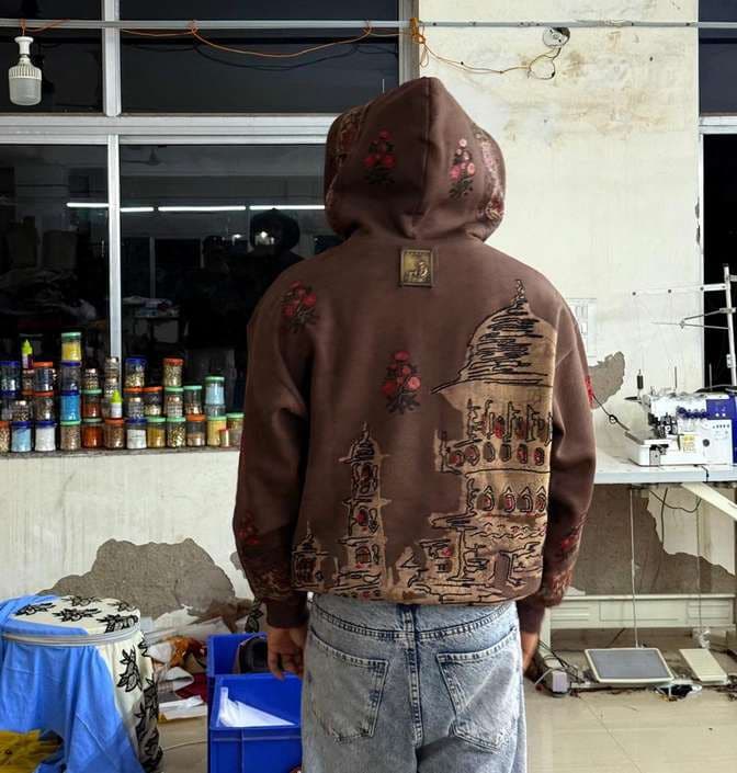 The back of a brown hoodie with an embroidered architectural scene and small red appliqué flowers.